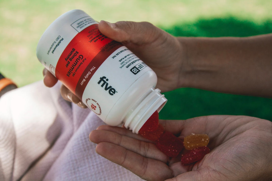Hand pouring red and orange gummies from a white bottle labeled 'five' with text 'Full Spectrum CBD Gummies 50mg per gummy' and 'The Daily Buzz'.