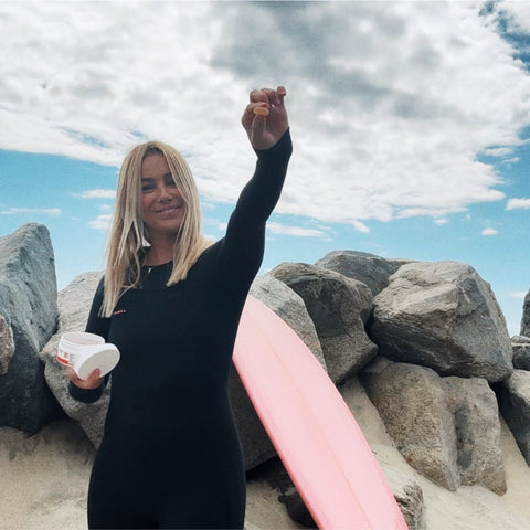 Woman in a black wetsuit holding a container and a small object, standing on a rocky beach with a pink surfboard leaning against rocks behind her under a cloudy sky.