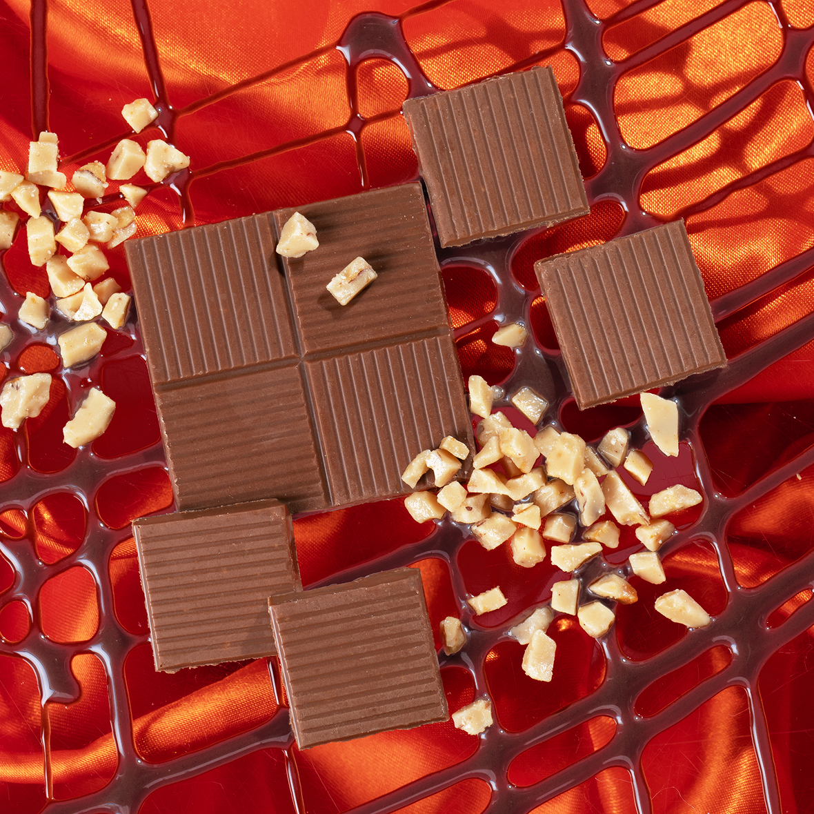 Chocolate squares and chopped nuts are arranged on vibrant orange fabric with a mesh-like design. The chocolates are neatly placed, some tilted, adding a sense of pattern to the composition.