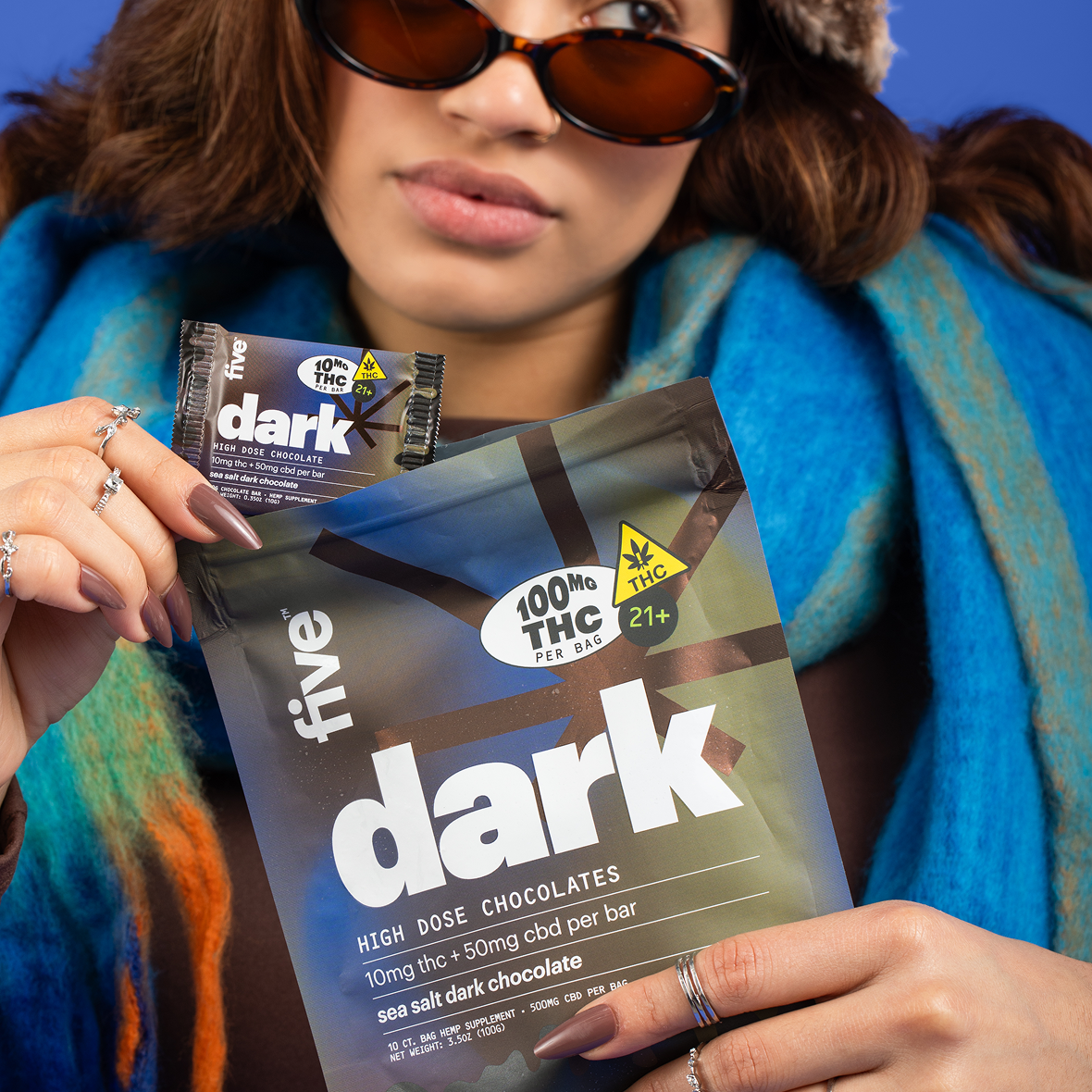 A person wearing sunglasses and a blue scarf holds a large package and a small packet of "dark" high dose chocolates. The packaging notes 100mg THC, with sea salt dark chocolate indicated.