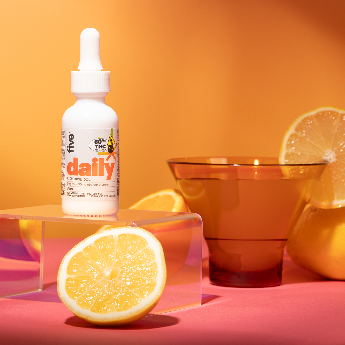 White bottle labeled five, daily, MICRODOSE OIL, 60MG THC, citrus; halved lemon and amber glass on orange.
