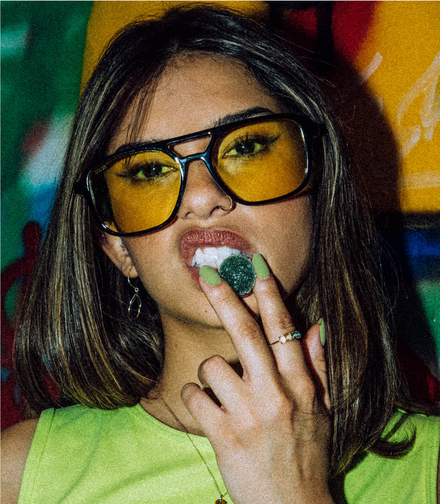 A woman in a green shirt eating a gummy with yellow tinted sunglasses.