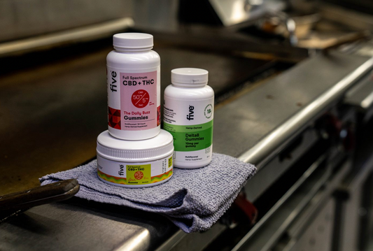 Front three-quarter view of three Five gummy containers showing CBD and THC and Delta8 product labels on a gray towel on a stainless steel counter