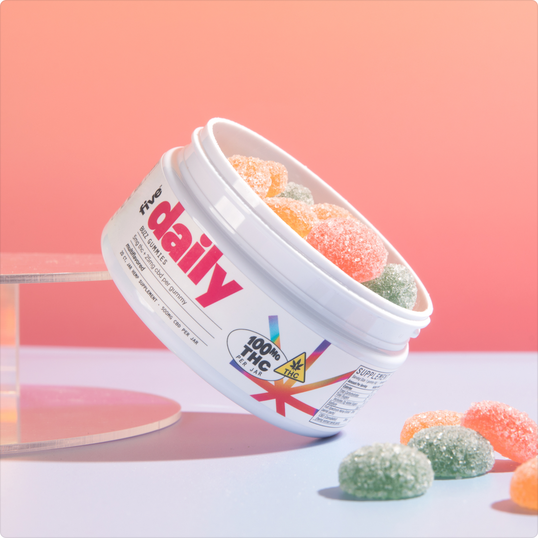 Angled view of Daily Buzz gummies jar showing sugar-coated orange and green gummies and 100 mg THC label on pink gradient background