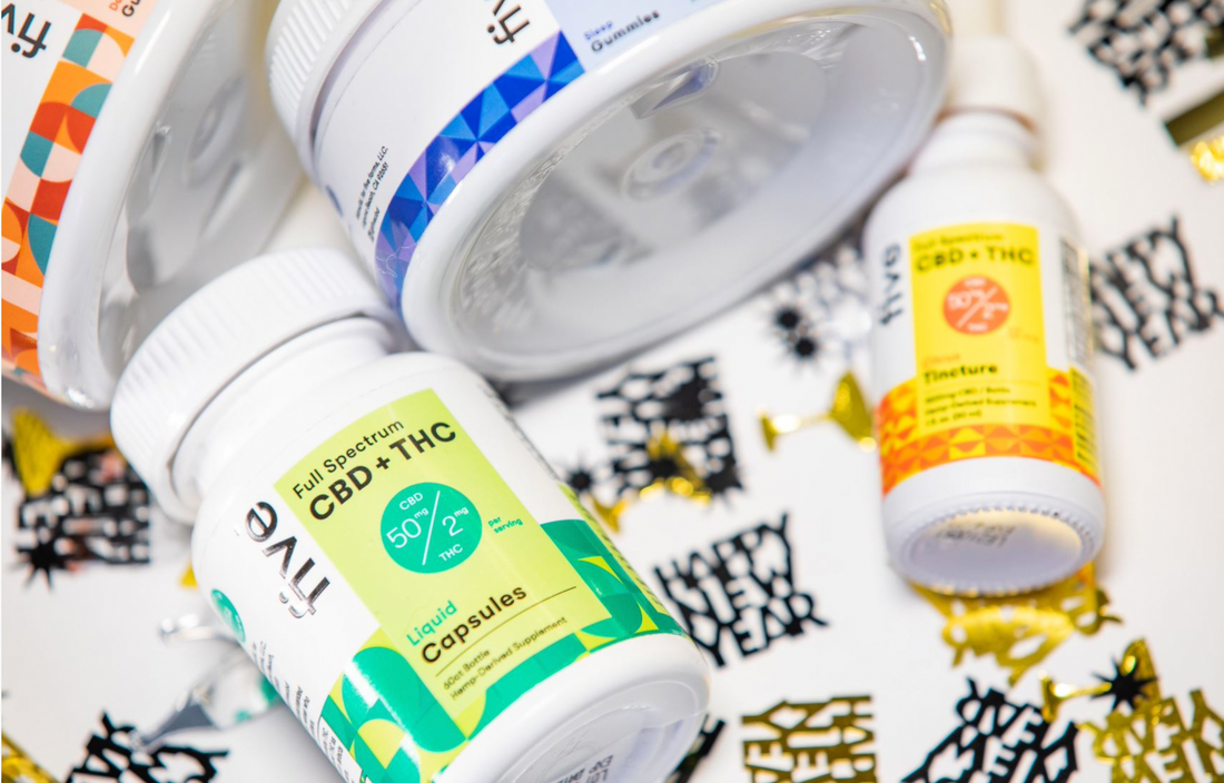 Bottles of CBD products labeled 'Full Spectrum CBD + THC', '50 mg CBD', '2 mg THC', 'Liquid Capsules', and 'Tincture' on a surface decorated with 'Happy New Year' confetti.