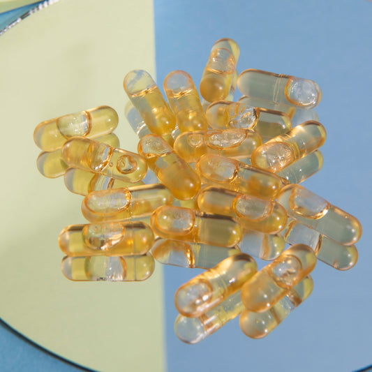 Yellow translucent softgel capsules with oil, scattered on a round mirror against pale blue and yellow background.