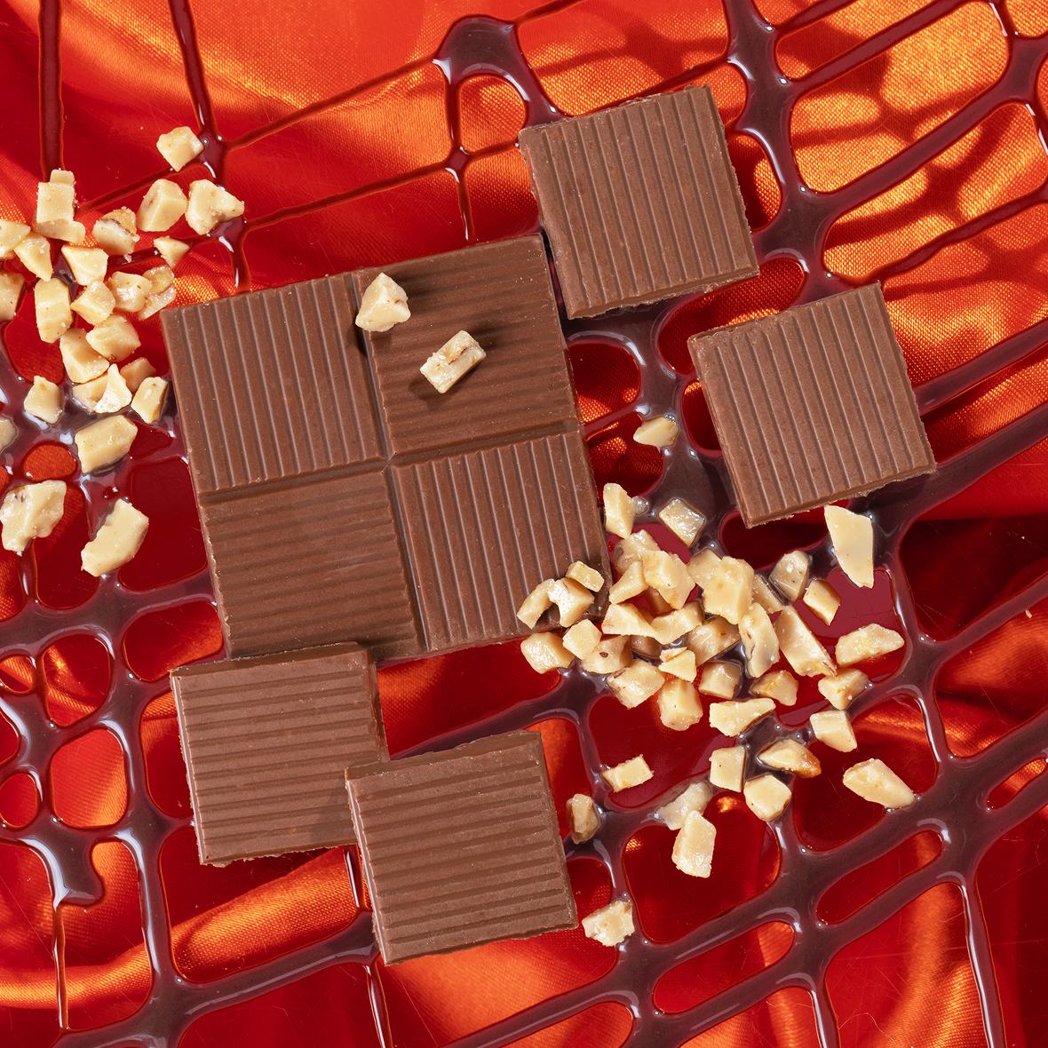 Chocolate squares and chopped nuts are arranged on vibrant orange fabric with a mesh-like design. The chocolates are neatly placed, some tilted, adding a sense of pattern to the composition.
