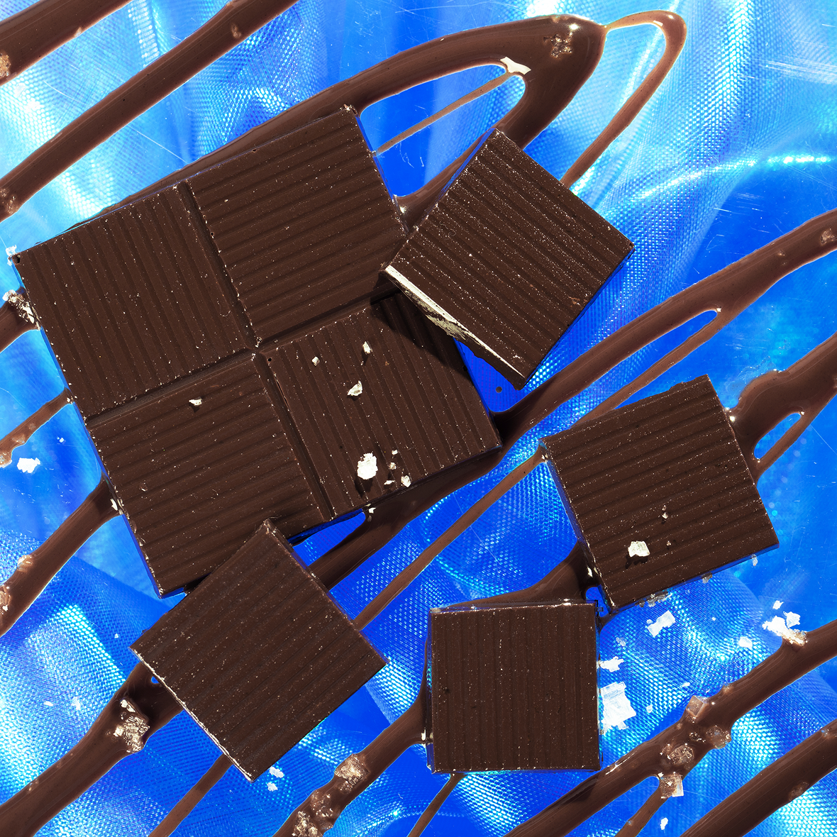 Squares of dark chocolate arranged on a blue surface with artistic drizzle patterns. Some chocolate pieces are broken, revealing the texture inside.