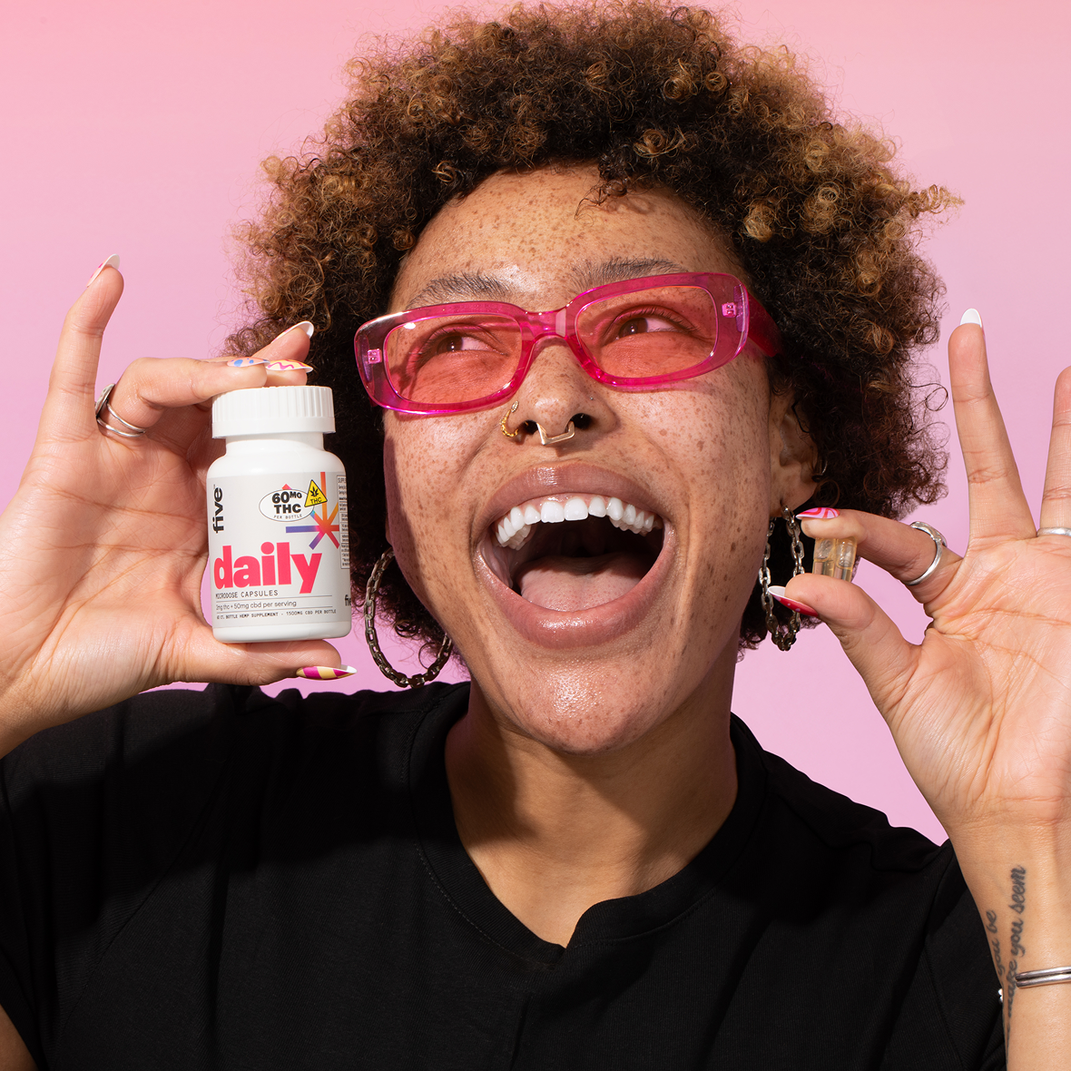 A person with curly hair and facial piercings is smiling and holding a bottle labeled "daily" in one hand and a small capsule in the other. They wear pink glasses and a black shirt against a pink background.