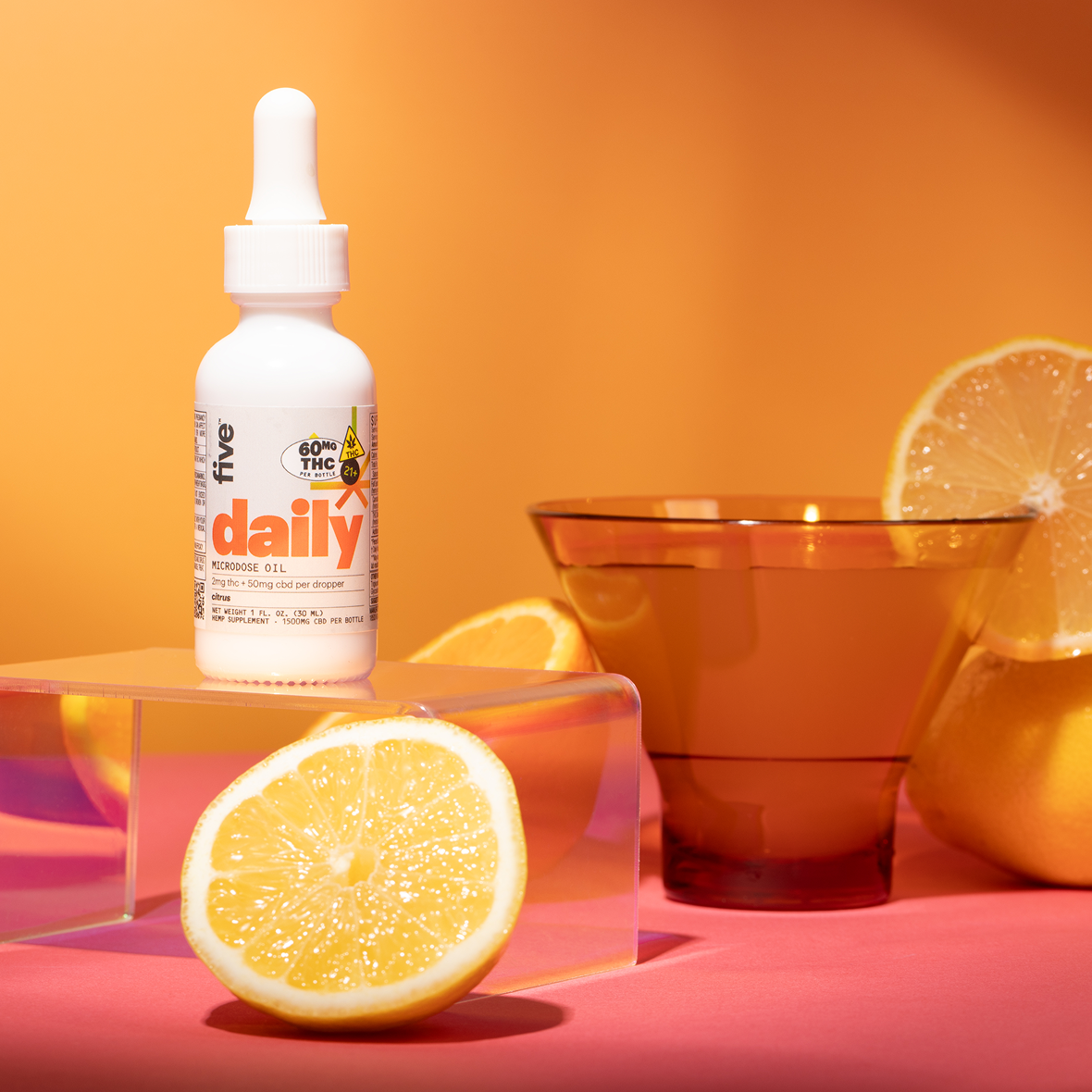 A white dropper bottle labeled "Daily" stands on a clear acrylic block. It is surrounded by lemon halves and an orange-toned glass on a pink surface with an orange background, suggesting a citrus-flavored product.