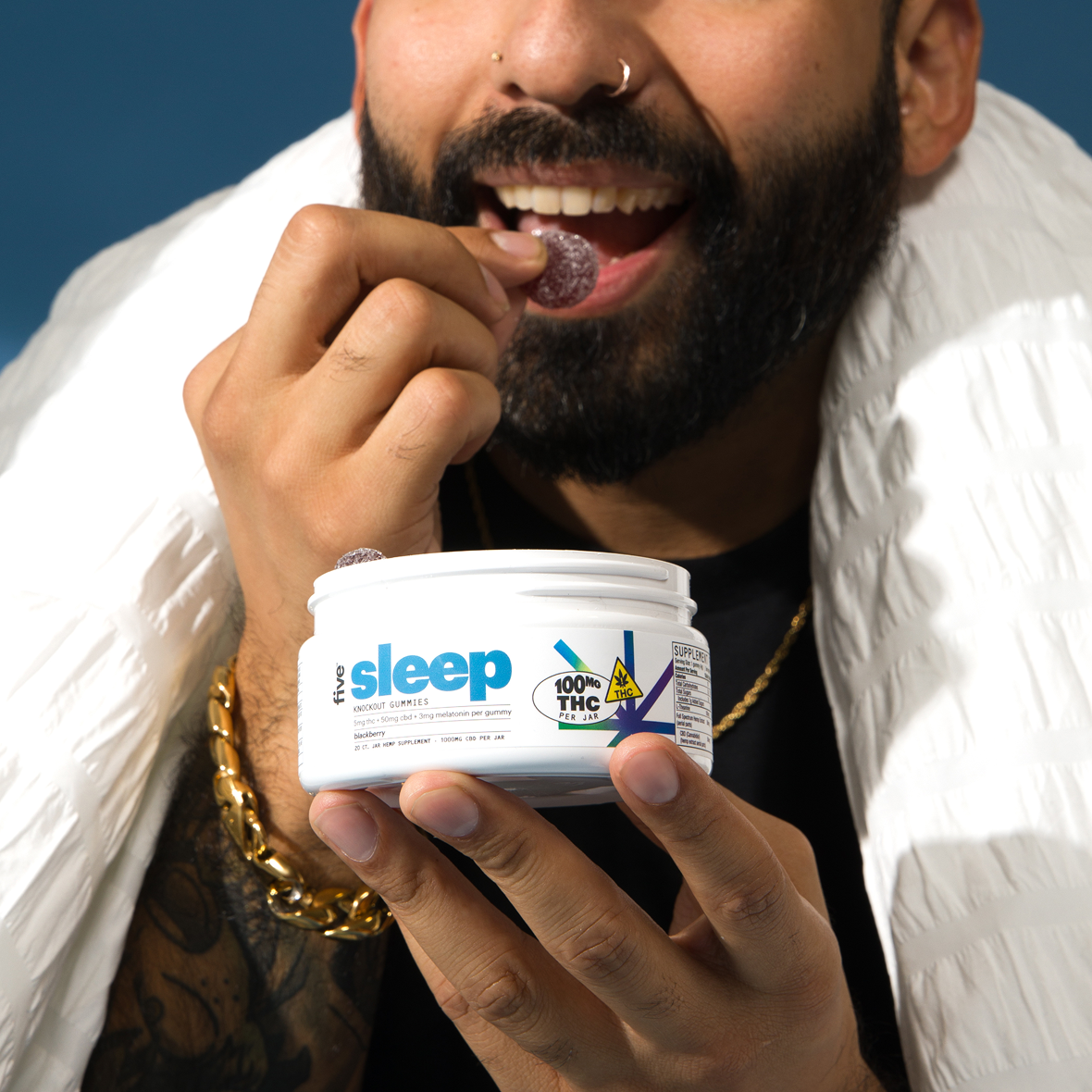 A person with a beard and nose ring, wearing a gold bracelet, is taking a gummy from a container labeled "sleep" with "100mg THC" on it. The person is smiling against a blue background, partially wrapped in a white blanket.