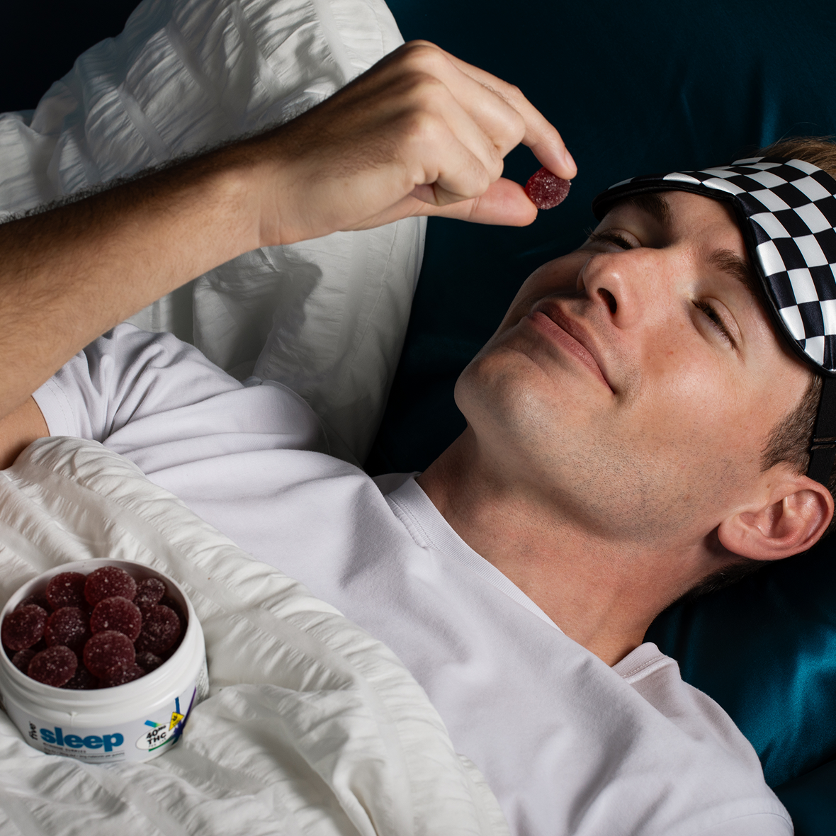 A person lying in bed with a checkered sleep mask on their forehead, holding a gummy above their mouth. A container of gummies is resting on the blanket beside them. They appear relaxed and ready to sleep.